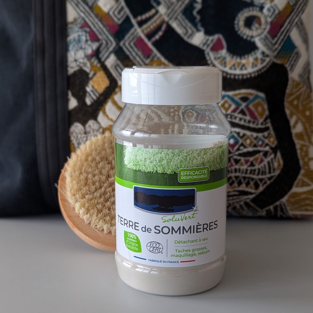 Un pot de Terre de Sommière et une brosse posés contre un sac en tissu jacquard